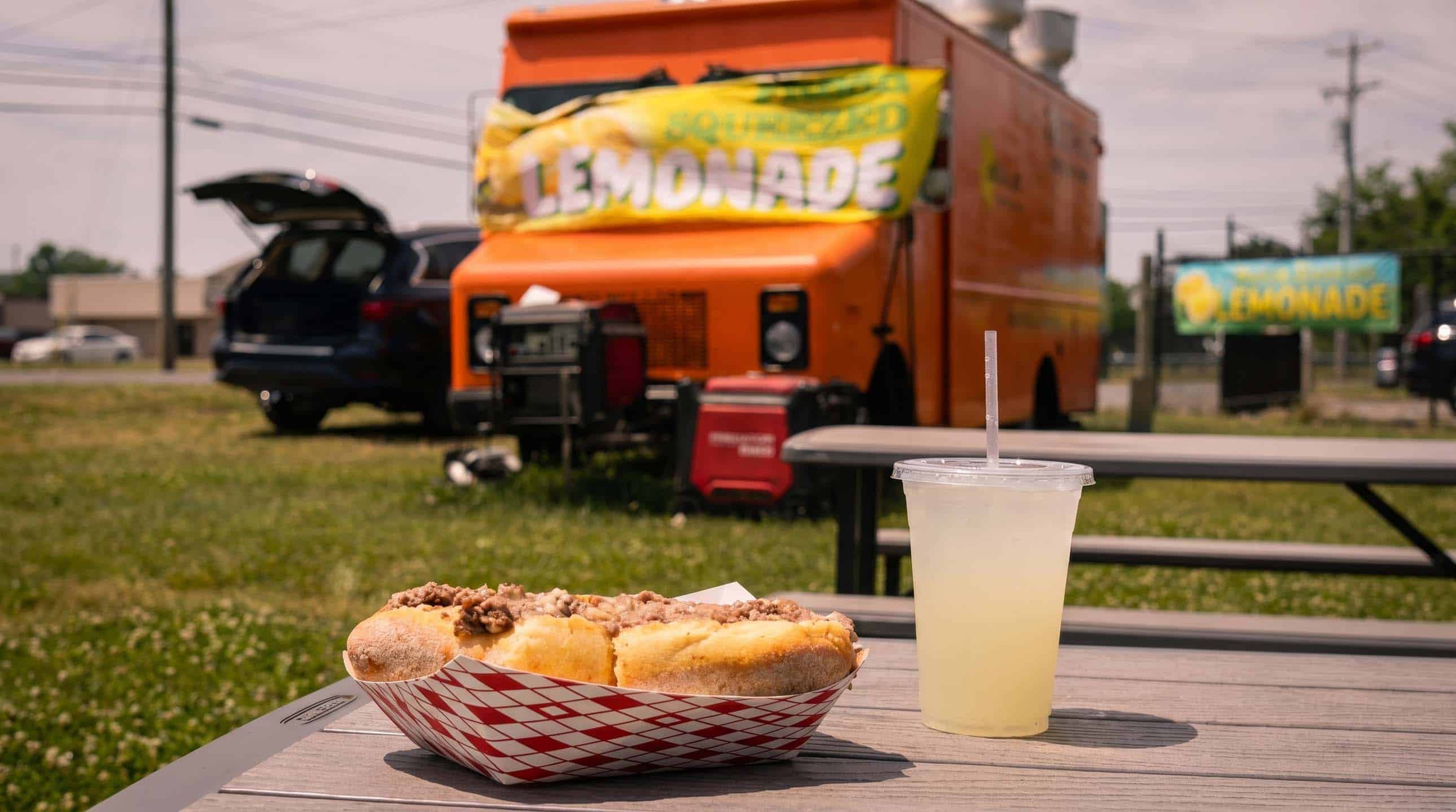 A boardwalk cheesesteak and a cup of fresh-squeezed lemonade on a picnic table in front of the orange Ahh la Cart food truck